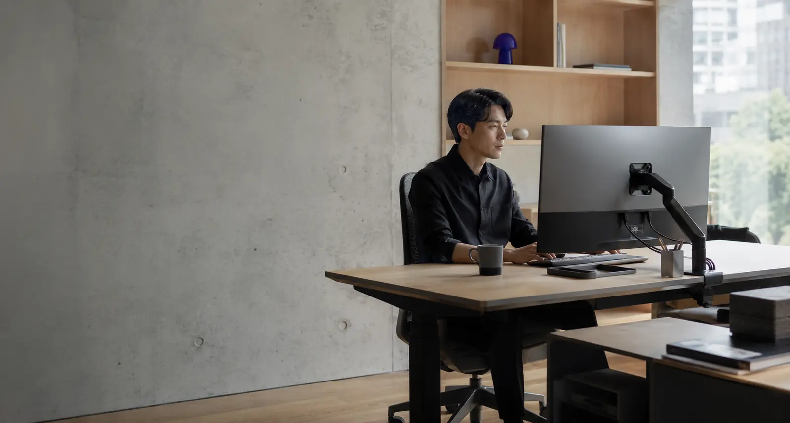 목재 책장 앞 전동 데스크에서 모니터 암과 함께 일하는 사람